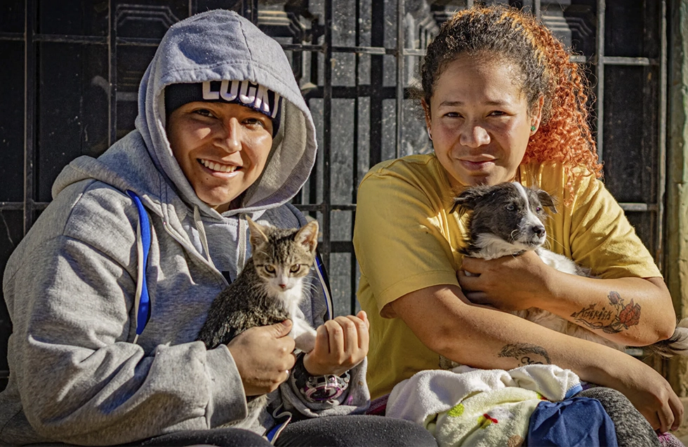 Two individuals smile warmly while holding their rescued cat and dog outside a home in Central America.